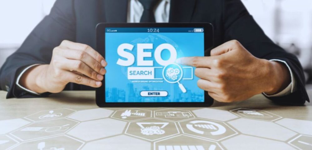 A business professional seated at a table holding a tablet that displays SEO SEARCH in bold text with a magnifying glass graphic, against a background of digital icons and hexagonal patterns.