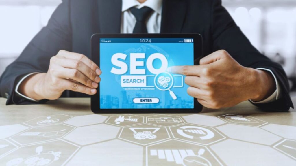 A business professional seated at a table holding a tablet that displays SEO SEARCH in bold text with a magnifying glass graphic, against a background of digital icons and hexagonal patterns.