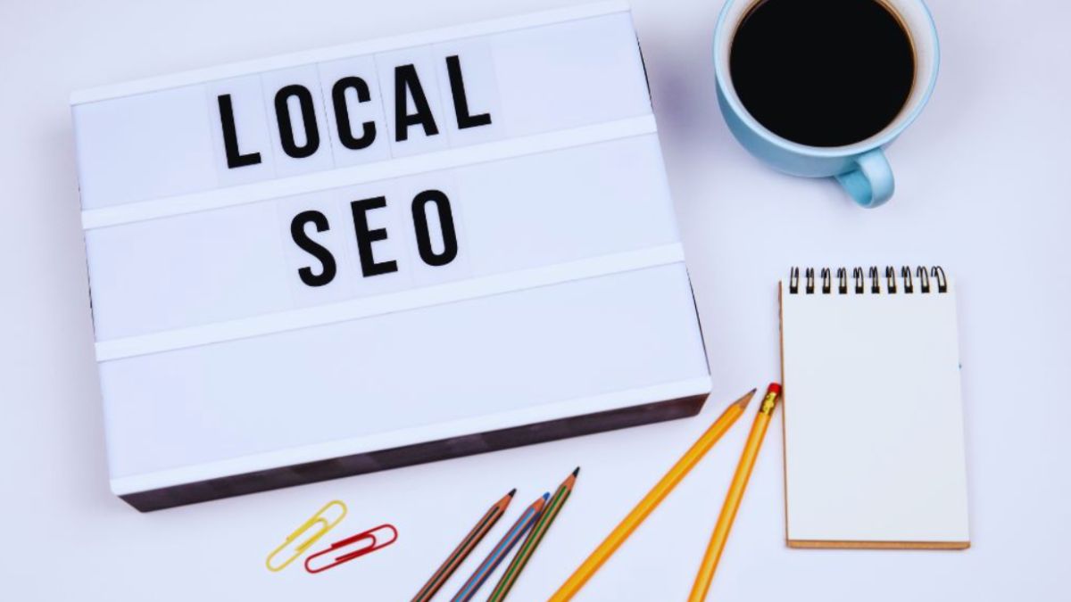 Flat lay of work items with a lightbox displaying LOCAL SEO in bold black letters, surrounded by pencils, clips, a notepad, and a coffee cup on a white background.