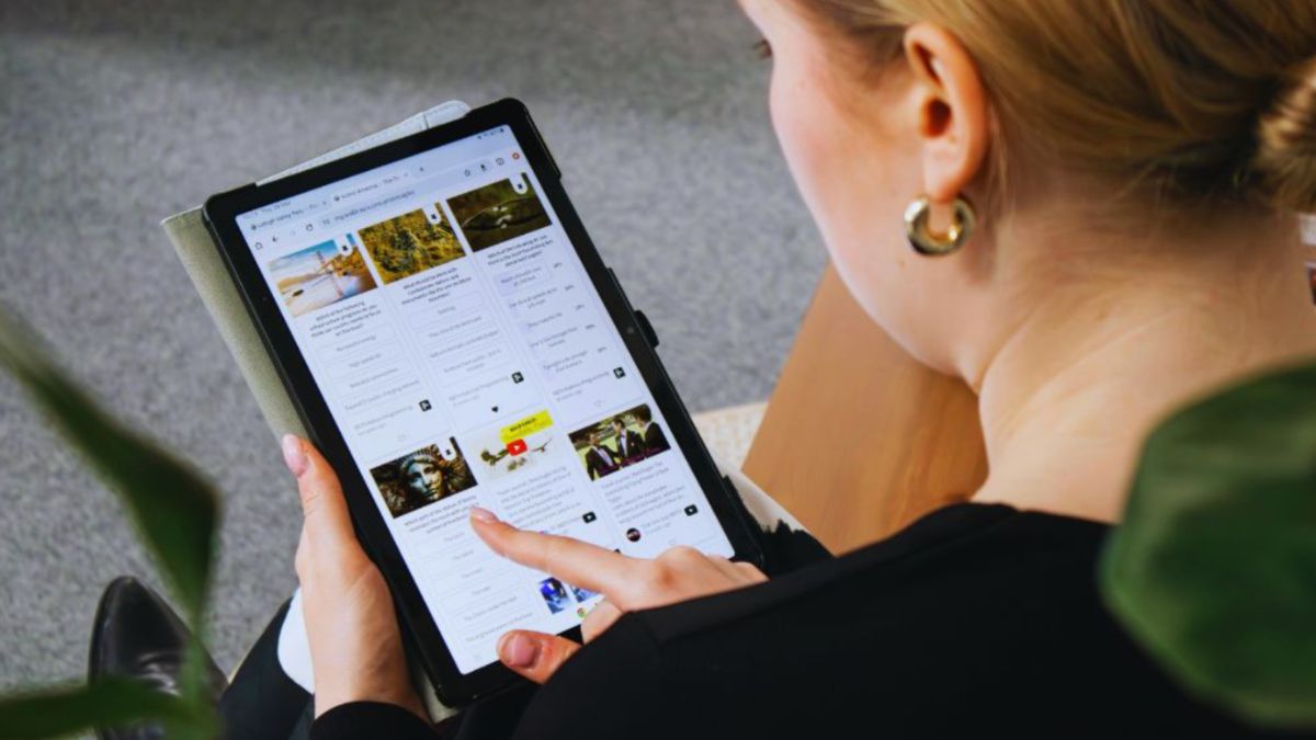 Person sitting and using a digital tablet to browse social media posts, selecting content for business promotion. The screen displays various post previews, highlighting online engagement and social media management for businesses.