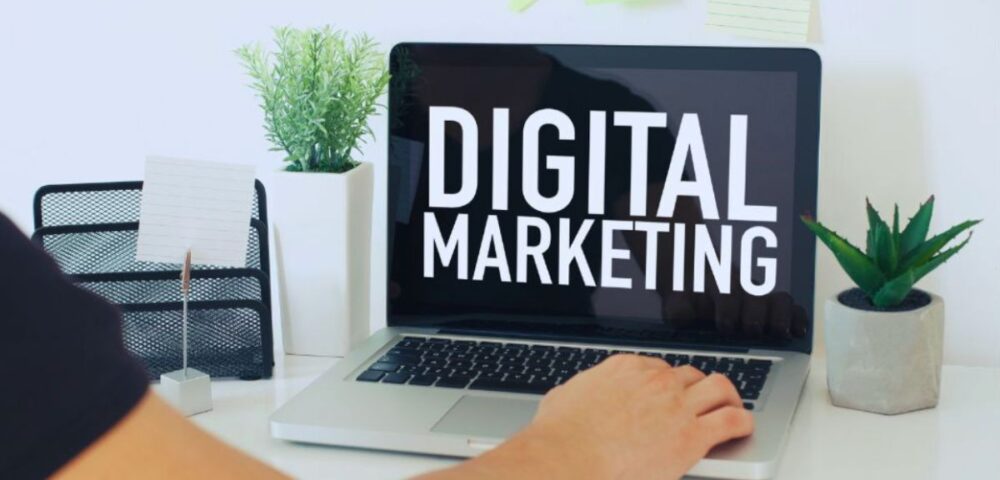 Person at a desk working on a laptop that displays “DIGITAL MARKETING,” with plants and office supplies on the workspace.