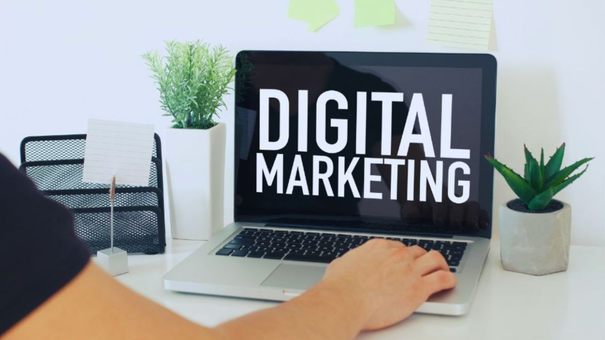 Person at a desk working on a laptop that displays “DIGITAL MARKETING,” with plants and office supplies on the workspace.