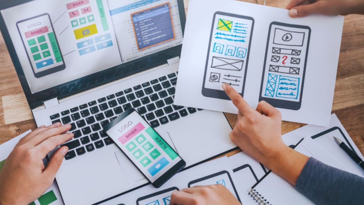 Hands collaborating on mobile app wireframes, with sketches of smartphone screens on paper and a laptop showing the same interface layouts, illustrating the web development process for a Palmetto Bay project.